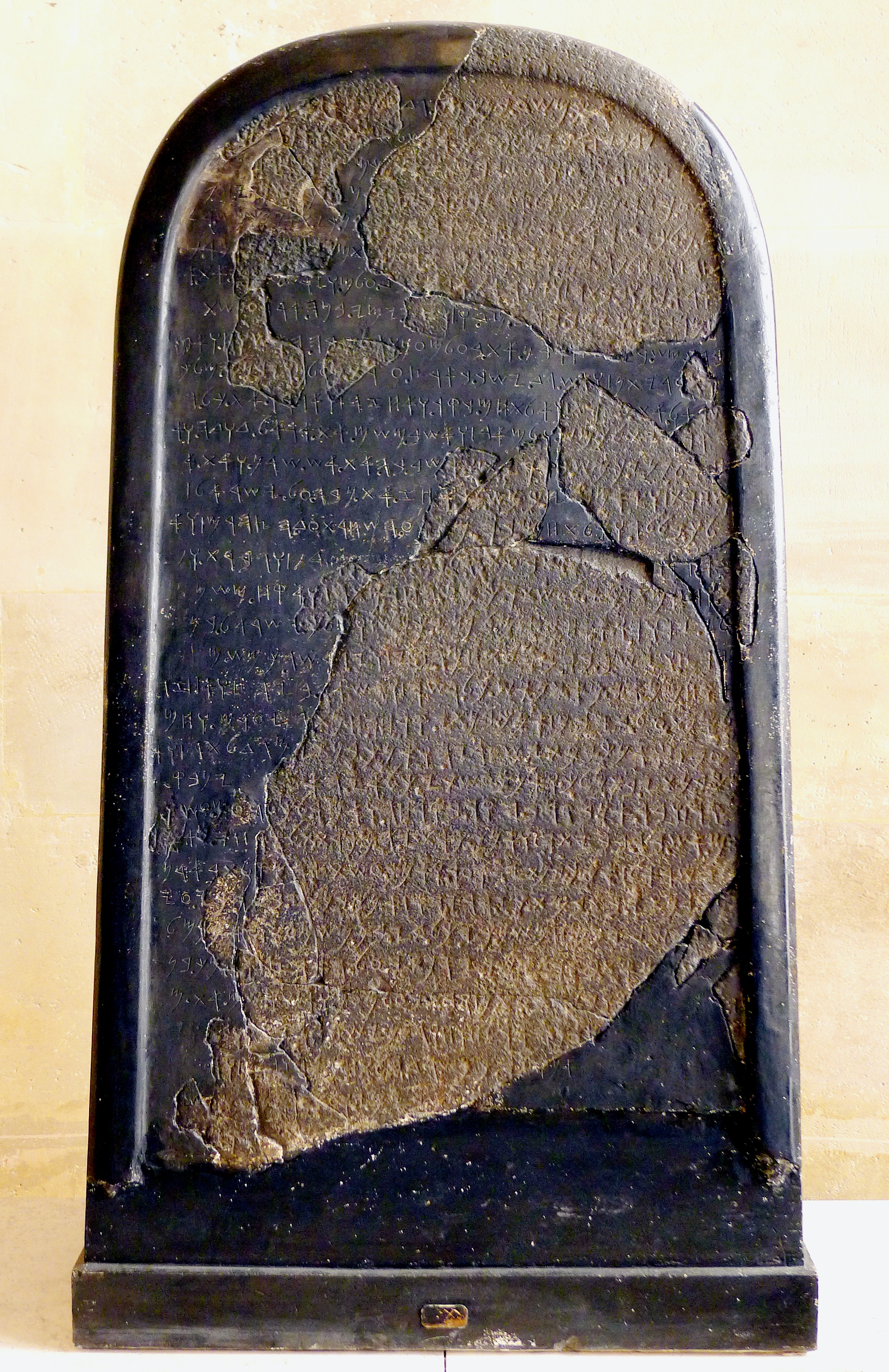 The Mesha Stele in the Louvre: a round-topped slab of black basalt, its rough inscribed fragments set into a smooth dark restoration, with rows of ancient letters running across the surface