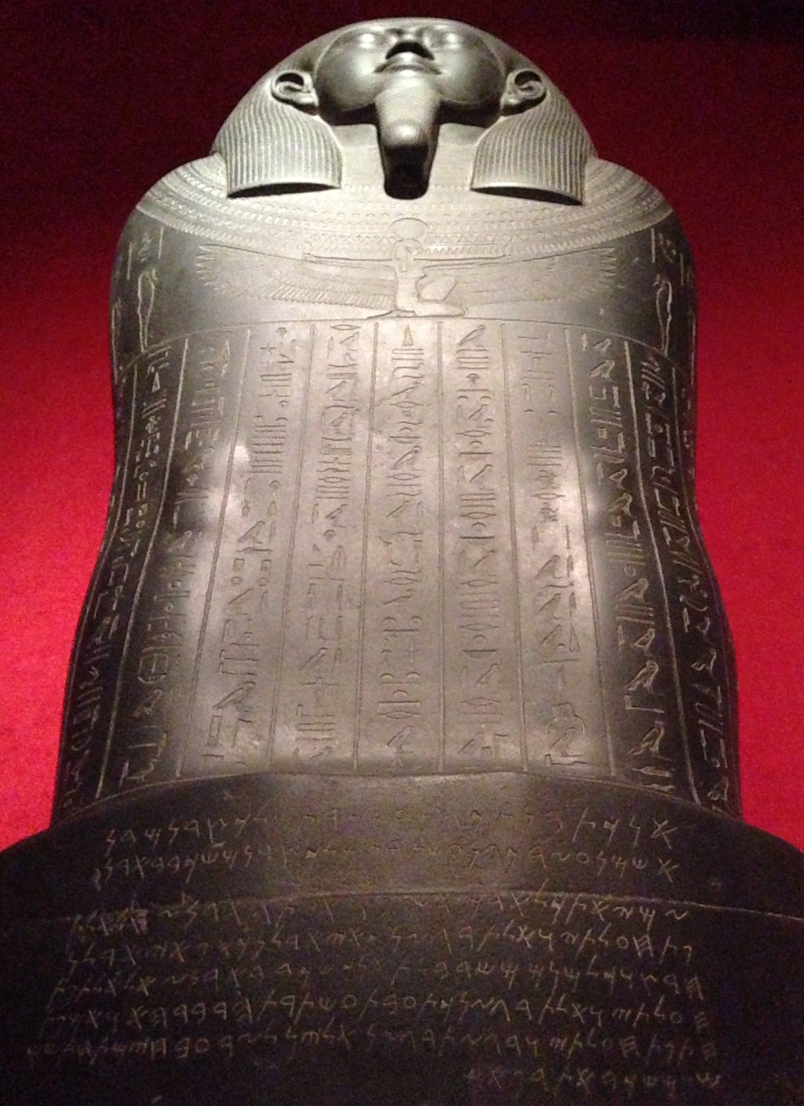 Tabnit’s black stone sarcophagus seen from its foot: an Egyptian face and wig at the head, columns of hieroglyphs down the body, and lines of Phoenician letters across the base